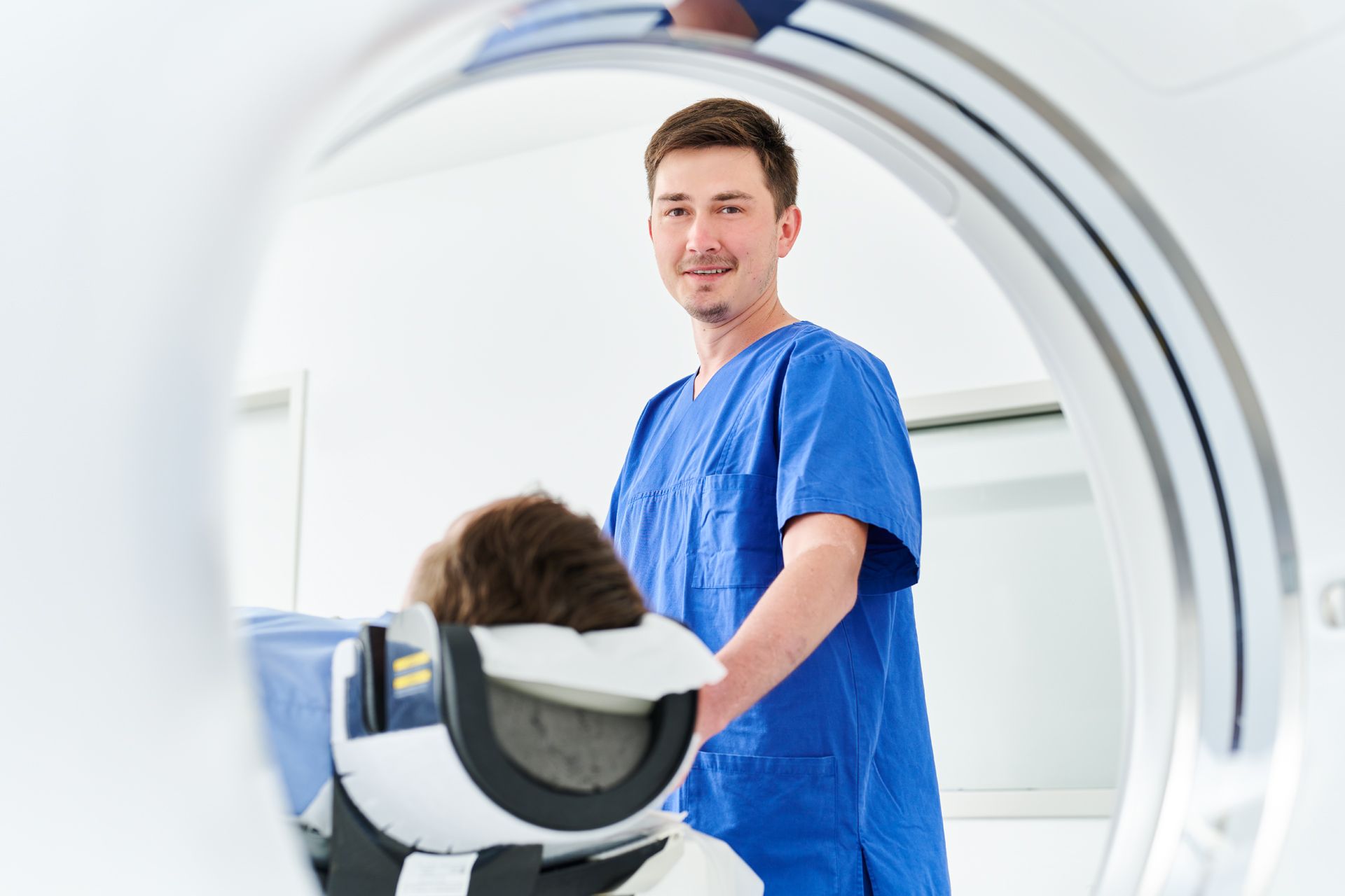 Blick von hinten durch eine CT-Röhre. Ein Patient liegt in einem CT und wird von Medizinischer Fachkraft betreut. 